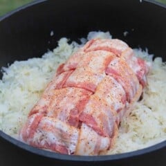 A raw pork loin wrapped in bacon sits nestled in sauerkraut in a Dutch oven.