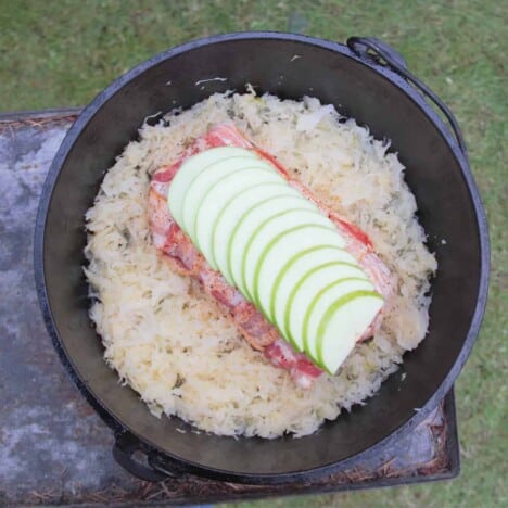 Looking directly down on a Dutch oven showing sauerkraut surrounding the bacon-wrapped pork loin topped with fanned out apple slices before being cooked.