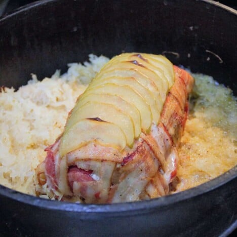 A cooked bacon-wrapped pork loin is topped with baked apples and surrounded by sauerkraut in a Dutch oven.
