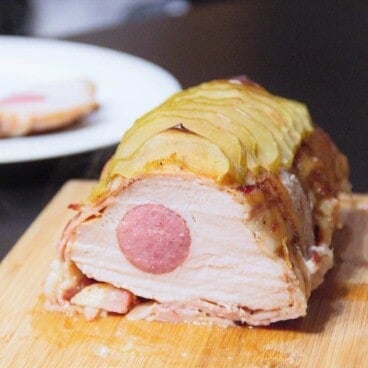 The roast is sitting on a chopping board sliced to expose the sausage within and the sliced apples on top.
