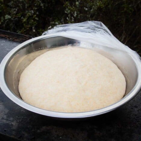 A stainless bowl with plastic wrap pulled back to expose the well risen whole wheat bead dough.