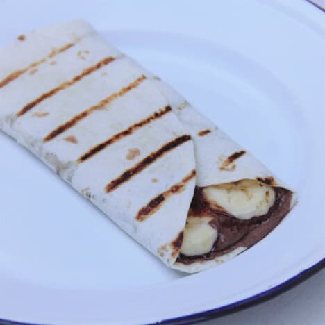 A grilled Nutella and banana wrap, with a bit of spread and bananas poking out of the edge, on a white camping plate.