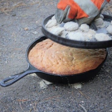 A Dutch oven lid with charcoal briquettes on it being removed from a skillet to expose the giant chocolate chip cookie inside.
