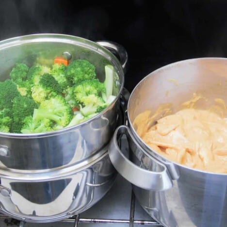 A camp gas stove with a steamer filled with vegetables and pot with chicken peanut sauce.