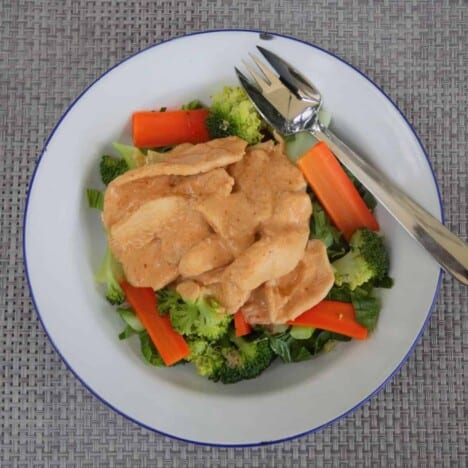 Looking down on a white camp plate with a blue edge on a serving of peanut chicken over steamed broccoli and carrots.
