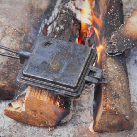 A square pie iron being held over campfire coals with the campfire flames in the background.