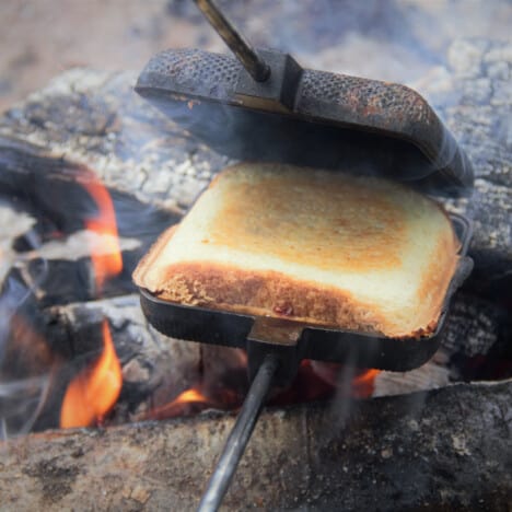 An open pie iron exposing the toastie inside, with the cooking campfire is in the background.
