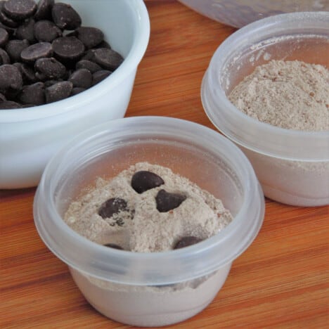 An individual plastic container of protein mix with chcolate chips, sitting next to one without and a large ramekin in the background full of chocolate chips.