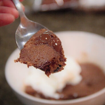 A spoonful of chocolate pudding in the foreground with the serving bowl of pudding with cream in the background.