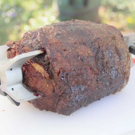 A rib eye roast still secured in the rotisserie spit resting on a white chopping board.