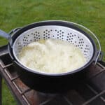 A cast iron deep fryer on a gas camp stove bubbling as it cooks apple fritters.