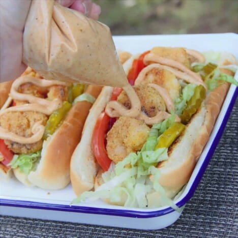A hand is squeezing remoulade sauce from a plastic bag over two finished po'boy sandwiches on a white camping plate.