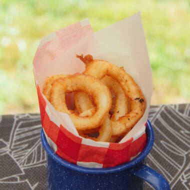 A blue tin cup sits on a picnic table with red and white paper rolled inside, filled with golden brown fried calamari rings.