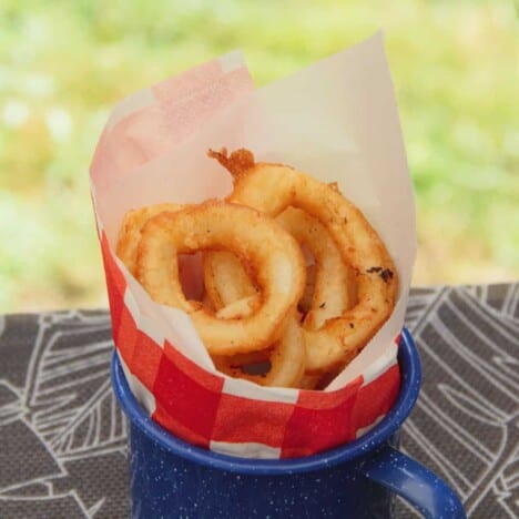 A blue tin cup sits on a picnic table with red and white paper rolled inside, filled with golden brown fried calamari rings.