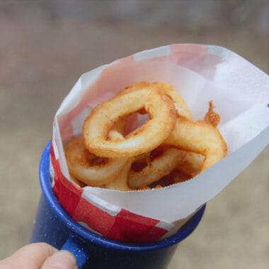 A pile of golden brown fried calamari rings sits in a red and white piece of paper in a blue tin cup.
