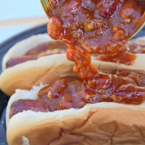 Chili is being poured on top of two hot dogs in buns on a black camping plate.