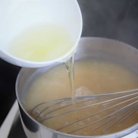 A white bowl with egg white in it being poured into the chicken and corn soup while being gently stirred with a whisk.