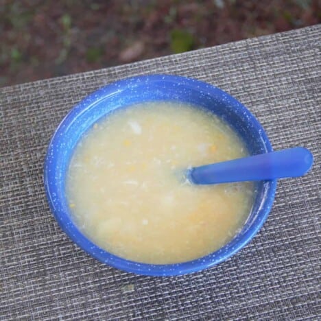 A serving of chicken and corn soup sits in a blue camping bowl with a blue spoon on a gray placemat.