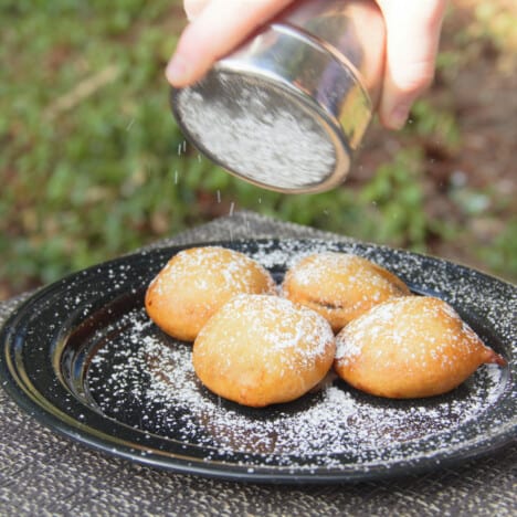 Four deep fried Oreos on a black camp plate being dusted with powdered sugar.