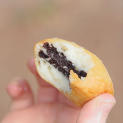 A hand holding a golden brown deep fried Oreo with a bite out of it exposing the cookie and puffed batter around it.