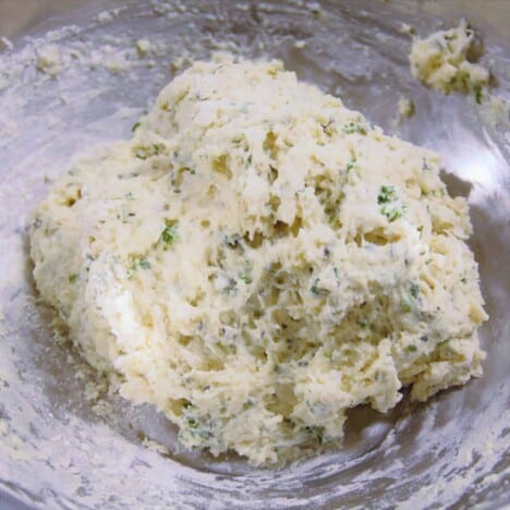 A ball of very wet dough speckled with green fresh herbs sits in a stainless steel camp bowl.