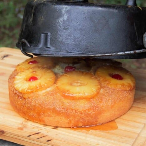 A hot pineapple upside down cake is being flipped out of a Dutch oven onto a wooden cutting board.