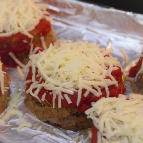 A close up shot of a single pizza mini meatloaf topped with pizza sauce and raw shredded cheese, on a foil-lined baking sheet.