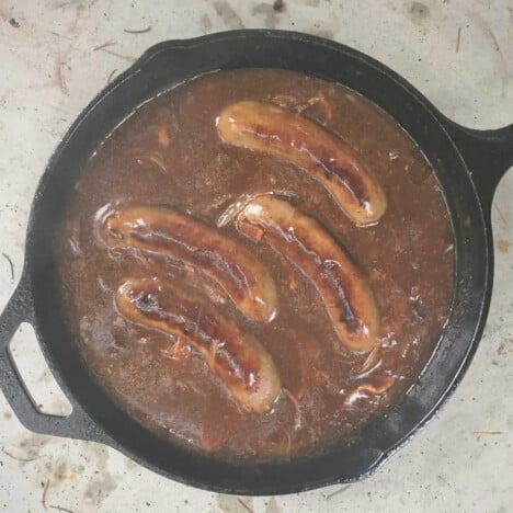 Looking down on a skillet with four sausages in an onion gravy.