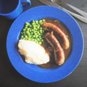 Looking down on a blue camp plate with a serving of sausages and gravy with a side of mashed potato and peas