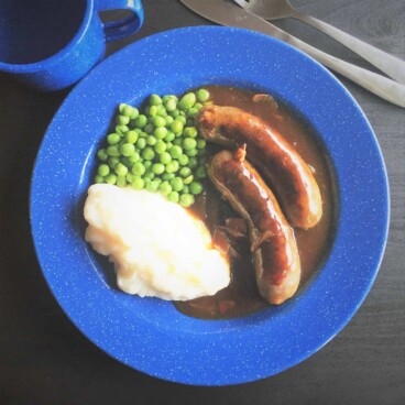 Looking down on a blue camp plate with a serving of sausages and gravy with a side of mashed potatoes and peas.