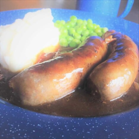 Close up of sausages with gravy on a blue camp plate with mashed potatoes and peas in the background.