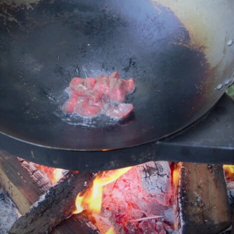 A large wok over a fire with the raw beef slices having just been added.