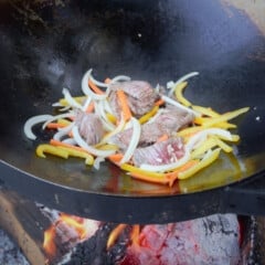 The beef, bell peppers, onions, and carrots cooking in a wok over a campfire.