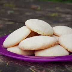 A purple plate holds a pile of light golden, round sugar cookies.