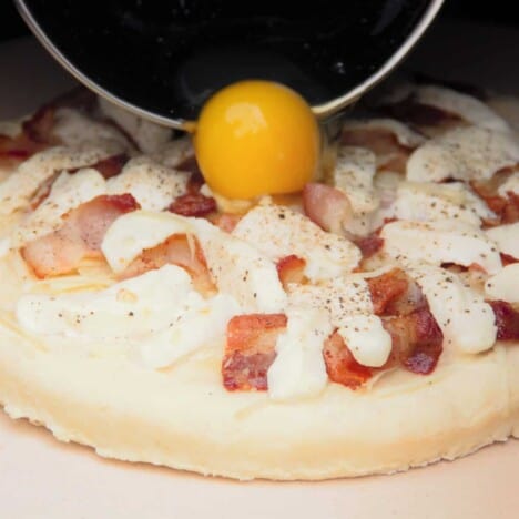 A raw egg being added to a bacon and egg pizza on a pizza stone at the beginning of the cook.