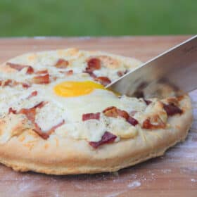 A bacon and egg breakfast pizza on a wooden chopping board being cut to serve.