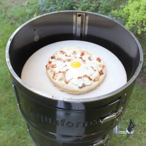 A cooked bacon and egg breakfast pizza sits on a pizza stone in a camp oven.