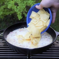 A skillet is simmering on a grill with a blue bowl of ravioli being poured in.