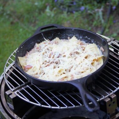A skillet over a grill filled with bacon and turkey ravioli having just been topped with parmesan