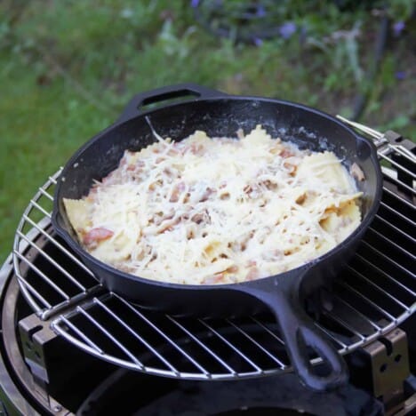 A skillet over a grill filled with bacon and turkey ravioli having just been topped with Parmesan cheese.