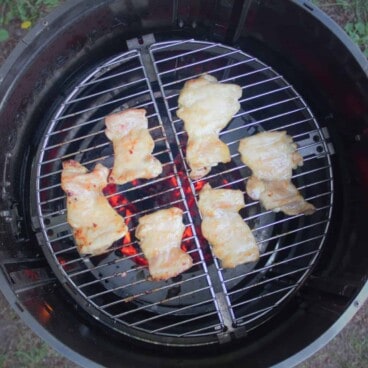 Looking down on six chicken thighs on a round grill over glowing red charcoals.
