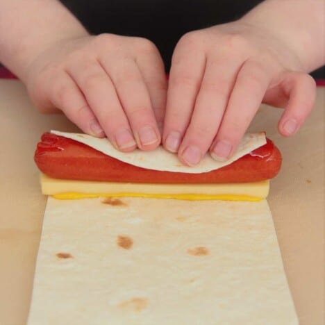 A young child rolls a hot dog and cheese slice in a tortilla on a work surface.