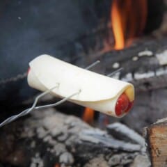 A Campfire hot dog wrap being held over a campfire coals on a toasting fork.