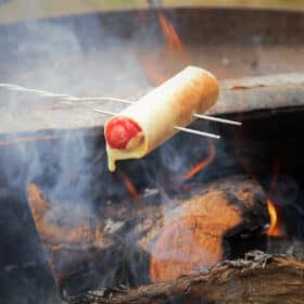 A hot dog wrapped in a tortilla with cheese oozing out being cooked over a campfire with a toasting fork