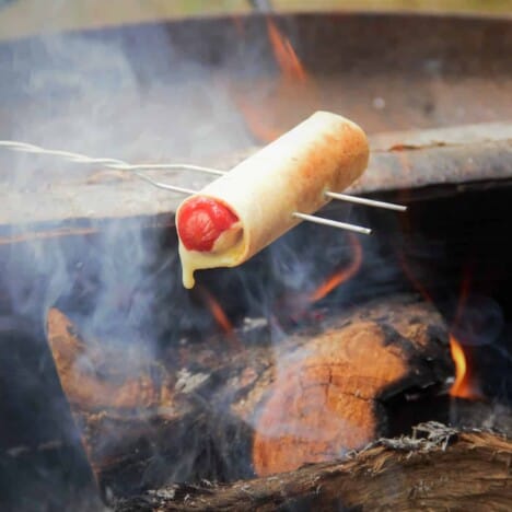 A hot dog wrapped in a tortilla with cheese oozing out being cooked over a campfire with a toasting fork.