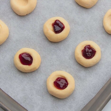 Several unbaked jam drop cookies, some with jam and some without, sit on a parchment-lined baking sheet.