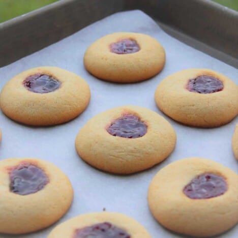 Several golden brown, baked thumbprint jam drop cookies sit on a parchment-lined baking sheet.