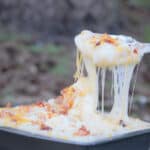The cooked loaded mashed potatoes in a square baking dish are having a spoonful being removed with many cheese strings back to the main dish.