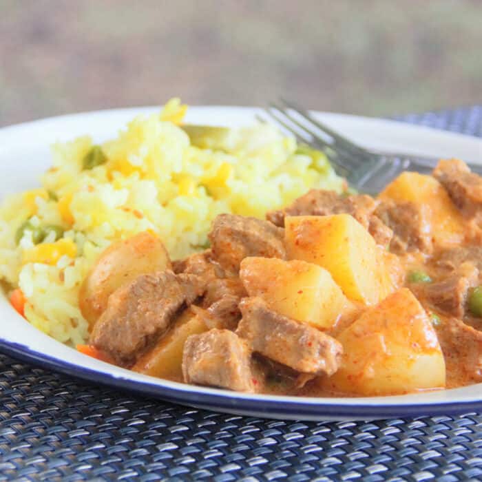 Looking across a white plate half filled with beef Massaman curry with large chunks of potatoes and yellow rice in the background.