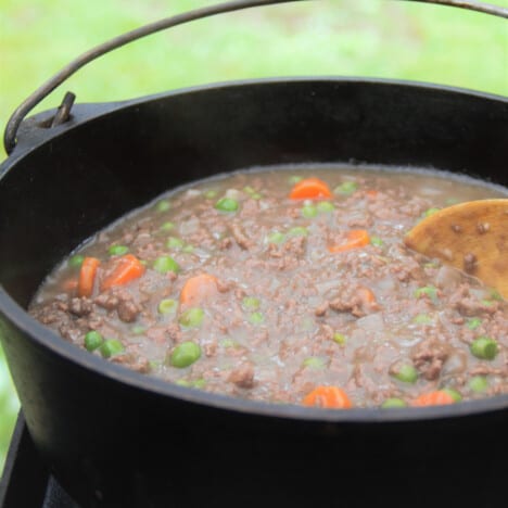 Looking into a Dutch oven while the base of ground beef, carrots, and peas simmers in a rich gravy.
